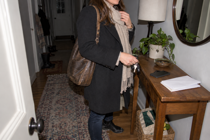 A woman just inside her front door, coat still on, holding her keys beside a face-down phone and mail on the hall table.