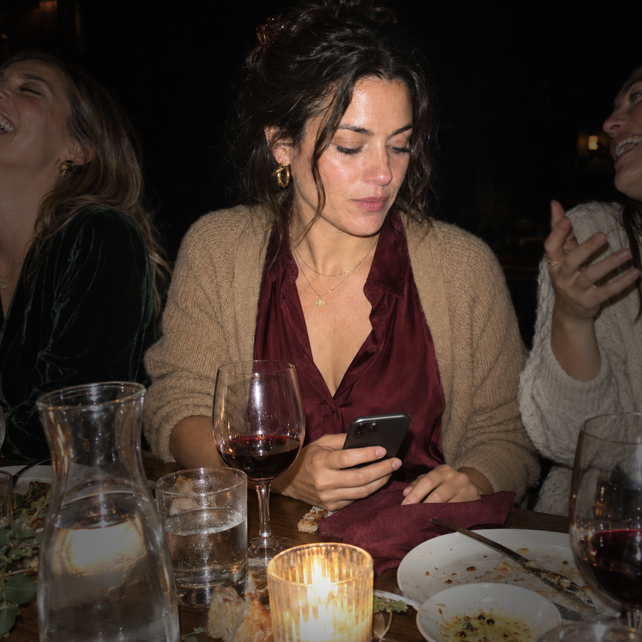 Three women at a restaurant table at night. The woman in the middle is still, eyes down on a phone held low, lips just closed, while the friends either side of her are mid-laugh.