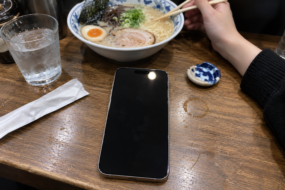 Looking down at a ramen counter, a phone face up with its screen dark, her hand lowering chopsticks into the bowl beyond it.